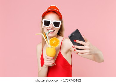 Smiling young woman girl in red one-piece swimsuit cap glasses isolated on pink background. People summer vacation rest lifestyle concept. Hold glass with cocktail doing selfie shot on mobile phone