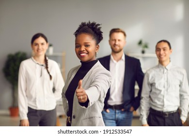 Smiling Young Woman Giving Thumbs-up Together With Diverse Multiracial People In Background. Good Business Idea, Excellent Choice, Positive Outcome, Winning Solution, Success, Job Satisfaction Concept