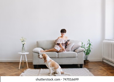 Smiling Young Woman In Glasses Looking With Smile At Beagle Dog Sitting On Carpet Beside Sofa. Indoor Portrait Of Lovely Brunette Girl Lies On Couch With Tablet While Her Puppy Resting On The Floor.