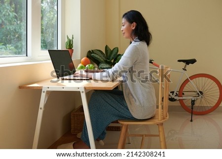 Smiling young woman with good posture wearing domestic clothes when working on laptop at home