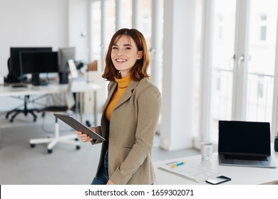 Smiling young woman in grey blazer and yellow sweater, with tablet pc in her hand standing half-turn and looking at camera in bright office interior - Shutterstock ID 1659302071