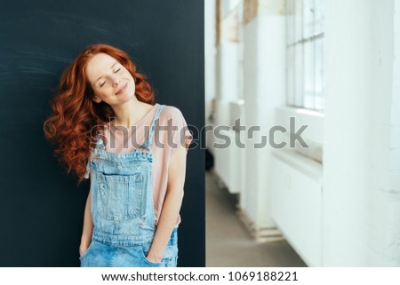 Smiling young woman with a happy serene smile wearing trendy denim dungarees standing with head tilted to the side indoors at home