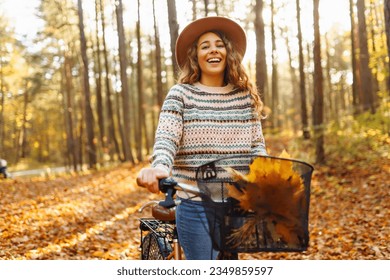 Smiling young woman in a hat and a stylish sweater with a bicycle walks and enjoys the autumn weather in the forest, among the yellow leaves at sunset.