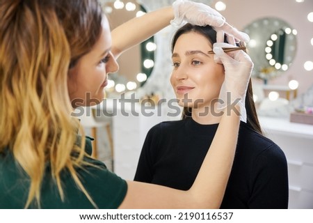 Smiling young woman having eyebrow correction procedure in beauty salon. Female esthetician in sterile gloves shaping woman eyebrows with cosmetic tweezers. ストックフォト © 