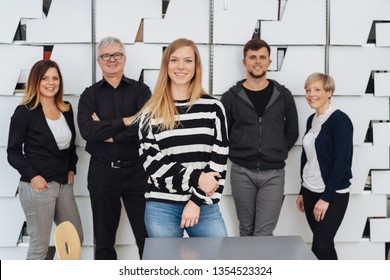 Smiling Young Woman With Her Business Team In Casual Clothing, Posing To Group Photo In Their Office, Standing Against Decorative White Wall And Smiling