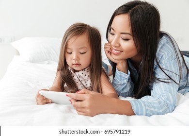 Smiling Young Woman And Her Little Daughter Looking At Mobile Phone While Relaxing Together In Bed At Home