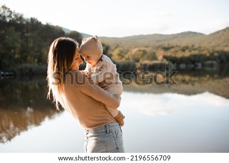 Smiling young woman holding playing with baby boy 1 year old wear knit clothes over nature background and lake with forest. Autumn season. Motherhood. 