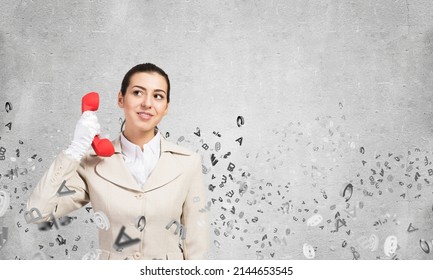 Smiling Young Woman Holding Retro Red Phone And Flying Various Letters. Call Center Operator In Business Suit With Phone. Hotline Telemarketing And Consultation. Business Assistance Service.