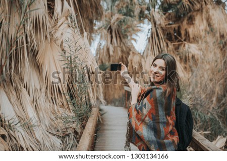 Smiling young woman holding smartphone, taking selfportraits outdoors, surrounded by palms.