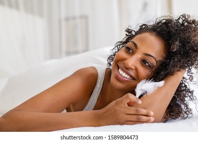 Smiling Young Woman Holding White Feather While Looking Away. Happy African American Girl Lying On Bed With Her Perfect And Soft Skin. Cheerful Dreamy Woman Feeling Good. Skincare And Beauty Routine.