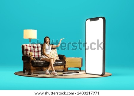 Smiling young woman in home wear sitting on couch near giant 3D model of mobile phone with empty screen over blue background. Online shopping, e-commerce. Mockup for text, ad, design, logo