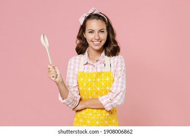 Smiling Young Woman Housewife 20s In Yellow Apron Checkered Shirt Hold Spoon Soup Ladle Dipper While Doing Housework Isolated On Pastel Pink Colour Background Studio Portrait. Housekeeping Concept