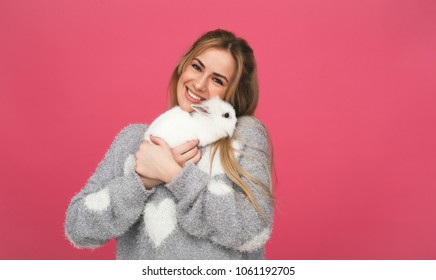 Smiling young woman hugs white rabbit. Pink background.