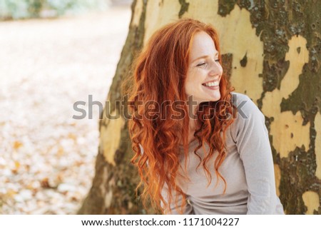 Smiling young woman leaning against the trunk of a tree with peeling bark looking to the side with a beaming smile of pleasure