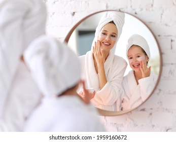 Smiling young woman and little daughter in white bathrobes and towels with eyes patches on faces looking at mirror during skincare procedure at home - Powered by Shutterstock