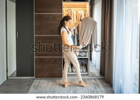 Smiling young woman with long curly dark hair in casual clothes standing in wardrobe and choosing outfit from hangers near shelves with various garments and shoes. High quality photo