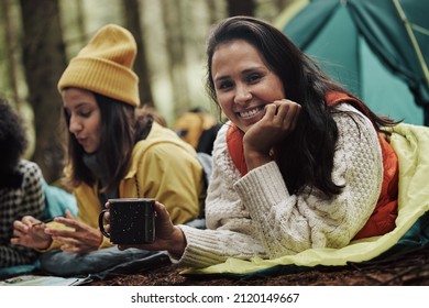 Smiling Young Woman Lying In Her Sleeping Bag And Drinking Coffee During A Camping Trip With Friends