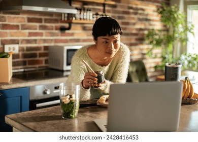 Smiling young woman making smoothie in the kitchen with laptop - Powered by Shutterstock