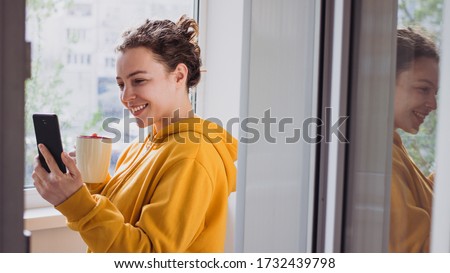 Smiling young woman making video call with smartphone near window balcony terrace at home. Caucasian millennial girl in yellow hoodie holding cup of coffee making selfie, sharing data on social 