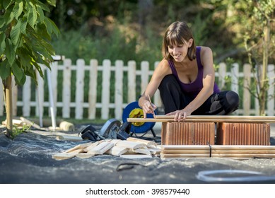 Smiling Young Woman Marking With A Pencil Where To Cut A Wooden Plank For A Backyard Fence In A DIY Concept.