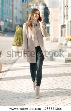 Smiling young woman with mobile phone walking in the sunny city street, girl wearing warm coat.