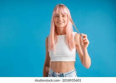 Smiling young woman with natural long pink dyed hair holding a strand of it and looking at camera, posing isolated over blue studio background. Beauty, hair care concept