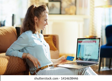 Smiling Young Woman With Opened On Laptop Online Travel & Booking Agency Site Holding Blue Credit Card At Modern Home In Sunny Summer Day. A Fake Website Is Made For Illustrative Purposes.