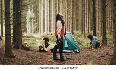 Smiling young woman in outdoor gear taking photos with a telephoto lens while camping with friends in the woods
