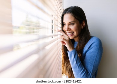 Smiling young woman peeking looking through venetian blinds on window at home - Powered by Shutterstock