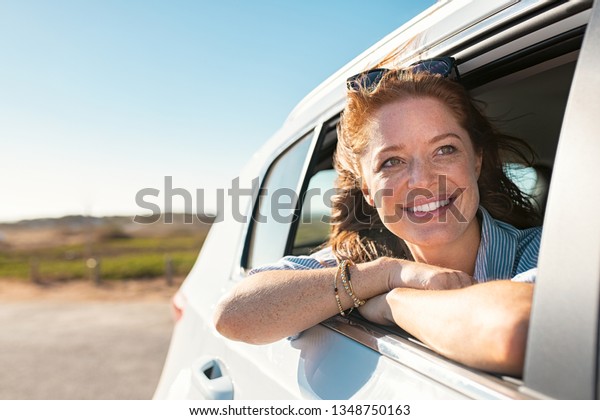 Smiling young woman peeking out of car window.\
Cheerful girl enjoying breeze from running car. Mature woman with\
red hair in the wind leans out of the car window and looking\
straight with copy\
space.