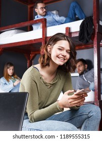Smiling young woman with phone in hands sitting on bed in hostel room