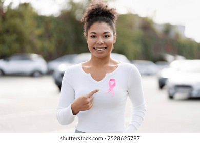 Smiling young woman pointing at pink ribbon attached to her shirt, supporting breast cancer awareness campaign - Powered by Shutterstock