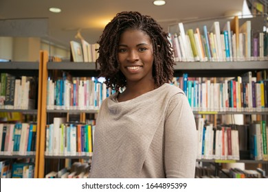 Smiling young woman posing at public library. Front view of cheerful lady with dreadlocks looking at camera. Knowledge concept