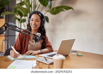 Smiling young woman recording podcast using microphone while laughing. Latin radio host working from home with copy space. Successful and cheerful girl taking notes while recording a podcast in studio - Powered by Shutterstock