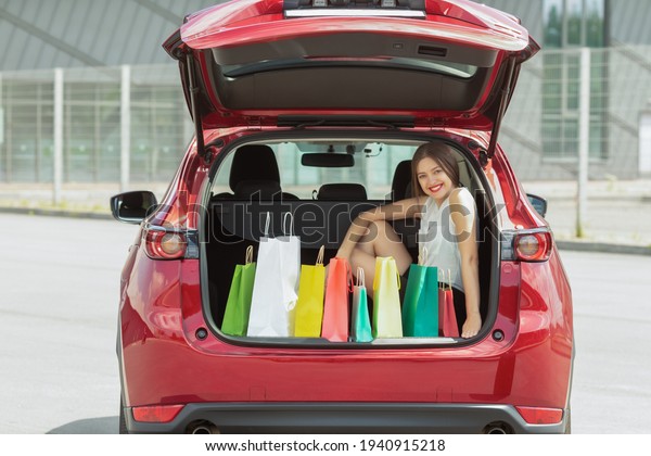Smiling  young woman in red car trunk with colourful\
paper bags. 