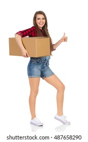 Smiling Young Woman In Red Lumberjack Shirt, Jeans Dungarees Shorts, And White Sneakers, Holding Box Under Her Arm And Showing Thumb Up. Full Length Studio Shot Isolated On White.