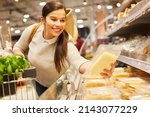 Smiling young woman at the refrigerated section in the supermarket buying cheese and reading shelf life information