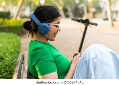 A smiling young woman relaxes on a park bench, enjoying music on her headphones, with an electric scooter beside her. - Powered by Shutterstock