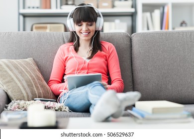 Smiling Young Woman Relaxing At Home On The Couch, She Is Wearing Headphones, Using A Digital Tablet And Taking Online Courses