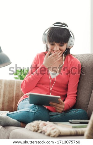 Smiling young woman relaxing at home on the couch, she is wearing headphones, using a digital tablet and watching a funny video online