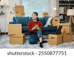 Smiling young woman relaxing on floor using smartphone surrounded by cardboard boxes, happy homeowner moving into new house