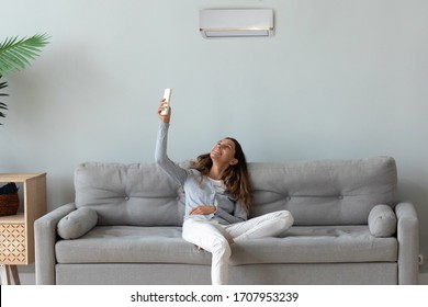 Smiling young woman relaxing on couch in living room, using air conditioner remote controller, switching, beautiful girl setting comfort temperature at home, enjoying fresh air, resting on sofa