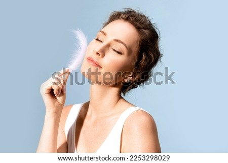 A smiling young woman runs a white feather across her smooth, delicate skin. Beautiful close-up portrait