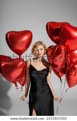 smiling young woman in satin slip dress standing with red heart-shaped balloons on grey