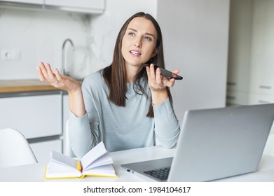 Smiling young woman is sending audio voice message by her smartphone working with laptop from home, female student using voice recognizable function for taking notes, searching information in network - Powered by Shutterstock