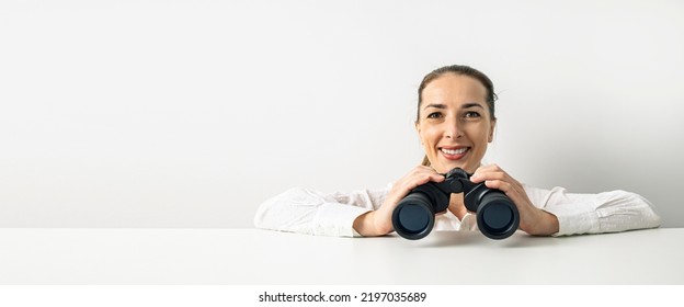 Smiling young woman in shirt with binoculars peeking out from behind cardboard. Banner. - Powered by Shutterstock