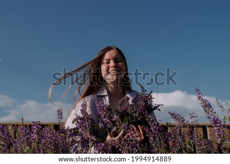 Smiling young woman in shirt holding bouquet of purple flowers standing alone among field in front wooden fence and blue sky enjoying of sunlight at golden hour. Summer outfit. Girl with flowers