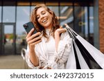 Smiling young woman shopper customer standing on city street holding shopping bags and smartphone. Sale ecommerce app. Consumerism, black Friday. Online shopping.