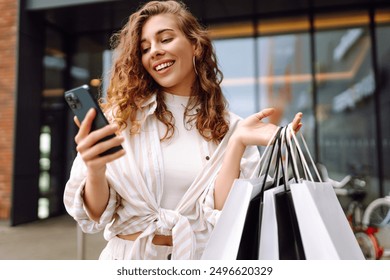 Smiling young woman shopper customer standing on city street holding shopping bags and smartphone. Sale ecommerce app. Consumerism, black Friday. Online shopping. - Powered by Shutterstock