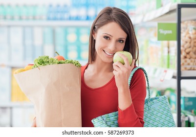 Smiling young woman shopping at the supermarket, she is holding a grocery bag with fresh organic vegetables